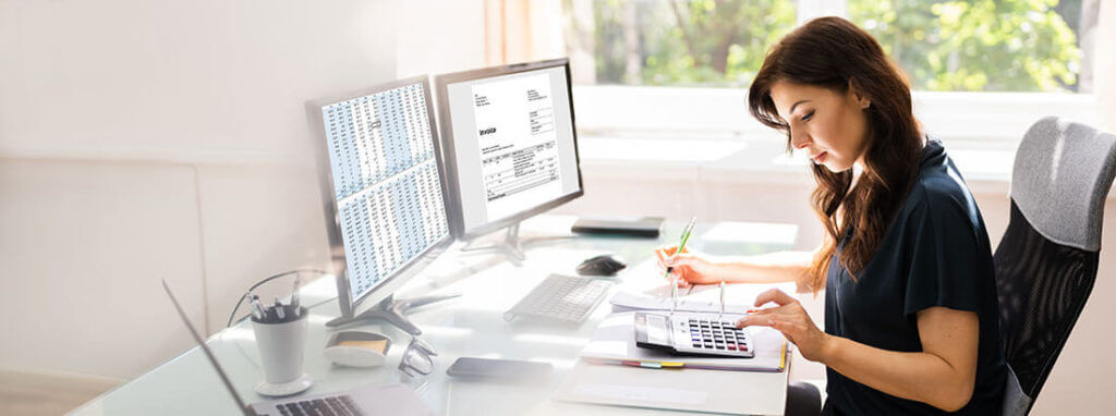 Eine Frau sitzt im Büro am Schreibtisch vor zwei Bildschirmen und tippt auf einen Taschenrechner während sie sich Notizen macht.