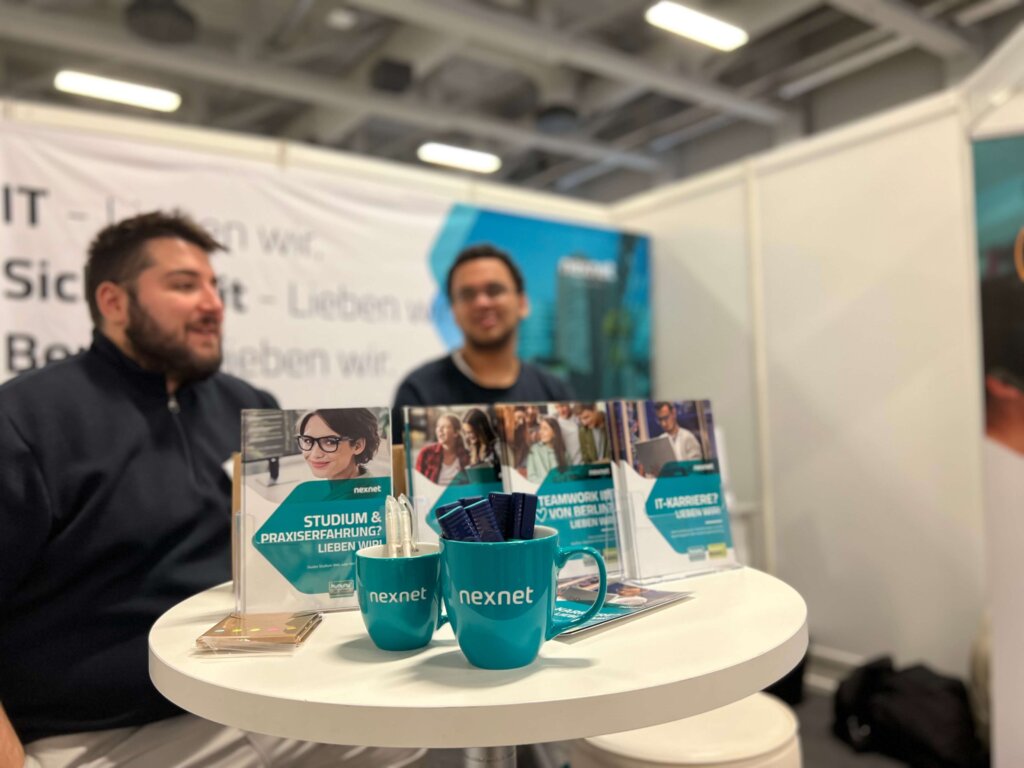 The nexnet booth at IT Berlin is shown here. Two trainees from nexnet can also be seen. You are standing in front of the table with the goodies.