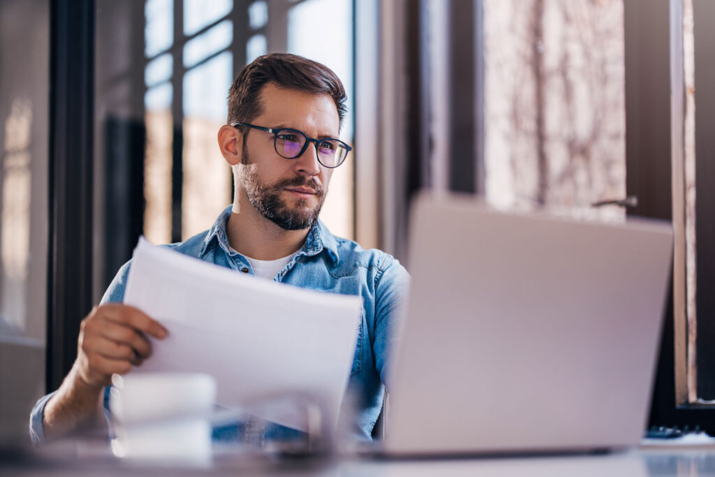 Abonnements verwalten mit nexnet.cloud, unserer Subscription Billing Lösung. Auf dem Bild ist ein Mann an einem Schreibtisch sitzend zu sehen. Er hält Dokumente in der Hand und schaut auf einen Laptop. Der Mann trägt eine Brille.