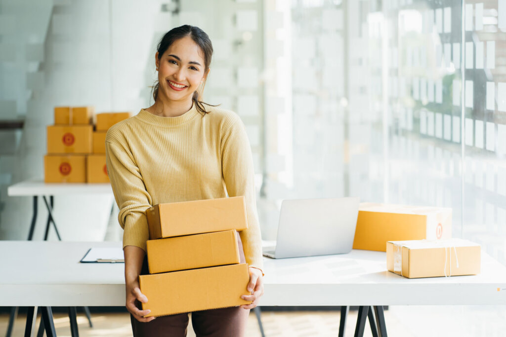 5 Tipps für den Start in den E-Commerce. Das Bild zeigt eine junge Unternehmerin, die drei Pakete in der Hand hält. Sie ist zur Kamera gewand. Sie steht in einem schlicht eingerichteten Büro.