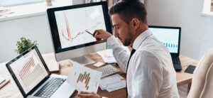 The picture shows a young man in an office. He is looking at some printed diagrams and comparing them with the images on the PC screen. The man is wearing a white shirt and tie.