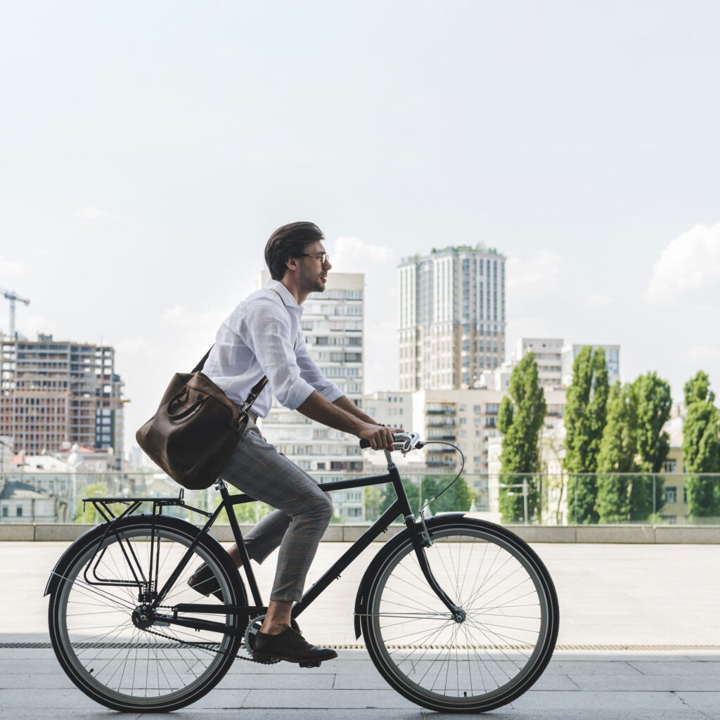 Auf dem Bild ist ein Radfahrer in sommerlicher Arbeitskleidung zu sehen. Der Radfahrer trägt eine große Umhängetasche. Er fährt auf seinem Fahrrad durch die Stadt.