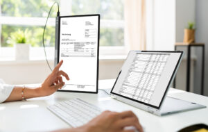The picture shows a person in an office, working at a desk and looking at two monitors. The monitors show tables and invoices. The person is making a payment reconciliation. Automatic payment reconciliation can be carried out with nexnet.