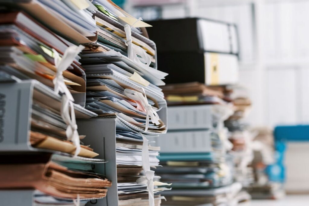 Das Bürokratieentlastungsgesetz IV ist da. Das Bild zeigt eine Nahaufnahme von diversen Papieren und Ordnern, aufgestapelt auf Tischen in einem Büro.