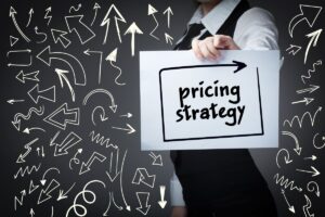 The picture shows a person holding up a piece of paper with pricing strategy written on it. In the background is a dark wall with many arrows painted on it, which look very different and point in all directions.