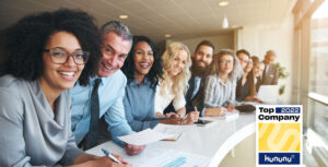 Many different young and old employees of a company are sitting at a long table. They are all looking happily at the camera. Slips of paper and diagrams are spread across the table. The Top Company 2022 award for nexnet can be seen in the bottom left-hand corner of the picture.