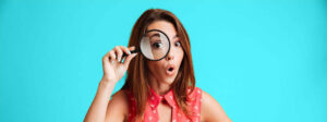 The picture shows a young woman looking through a magnifying glass. She is standing in front of a light blue background.