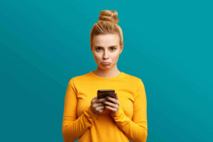 A young woman in a yellow shirt looks sadly at her hands, which she holds in front of her. She is standing in front of a light blue background.