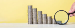 Stacks of coins are arranged in a row in steps. The largest on the left and the smallest on the right. A magnifying glass is pointed at the last, smallest stack of coins.