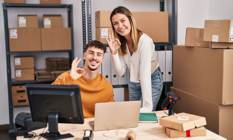 The picture shows an office with a wooden desk and metal shelves with various packages. A young man is sitting at a desk in front of a PC and smiling at the camera. Next to him is a young woman who is also smiling and looking at the camera. The man is wearing an orange sweater, the woman a white top and jeans. Both make an okay sign with their right hand.