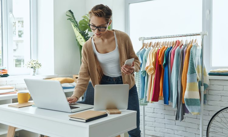 Subscription as a business model - nexnet's subscription billing solution makes this business model a success. The picture shows a young, modern businesswoman in a large room. She is standing at her desk and typing on her laptop. She is holding her cell phone in her other hand. In the background are items of clothing for sale.