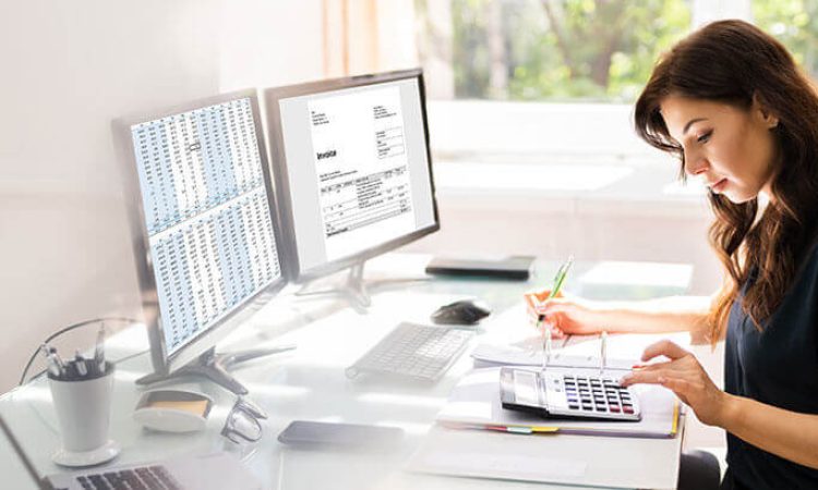Eine Frau sitzt im Büro am Schreibtisch vor zwei Bildschirmen und tippt auf einen Taschenrechner während sie sich Notizen macht.