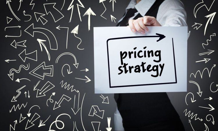 Das Bild zeigt eine Person, die einen Zettel hochhält, auf dem pricing strategy steht. Im Hintergrund ist eine dunkle Wand zu sehen, auf der viele Pfeile aufgemalt sind, die ganz unterschiedlich aussehen und in alle Richtungen verweisen.