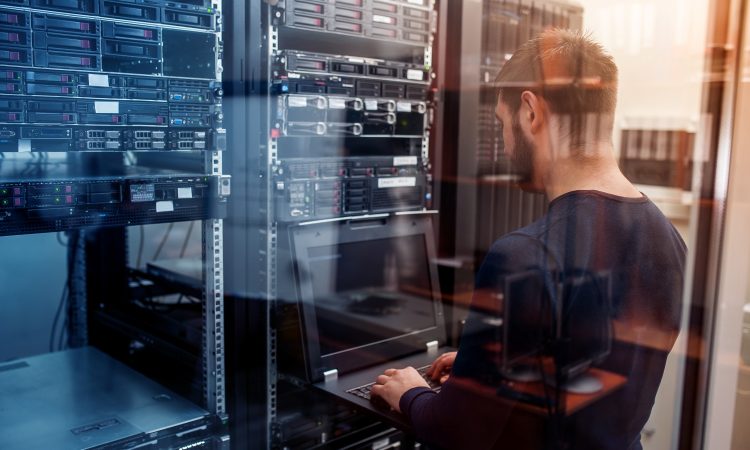 Das Bild zeigt einen Mann von hinten, wie er in einem Rechenzentrum steht und an einem Laptop etwas eintippt.