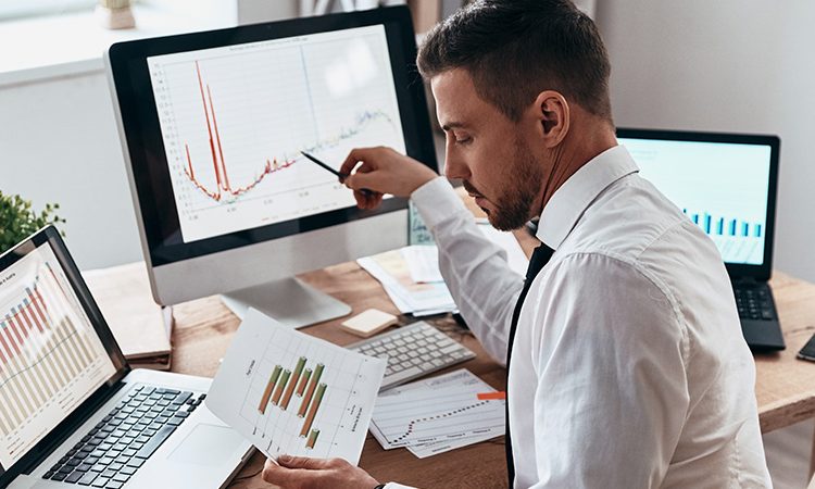 The picture shows a young man in an office. He is looking at some printed diagrams and comparing them with the images on the PC screen. The man is wearing a white shirt and tie.