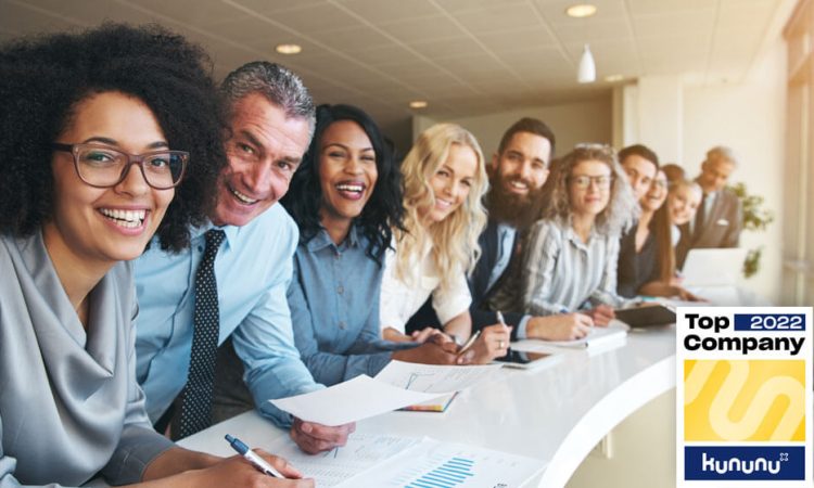 Many different young and old employees of a company are sitting at a long table. They are all looking happily at the camera. Slips of paper and diagrams are spread across the table. The Top Company 2022 award for nexnet can be seen in the bottom left-hand corner of the picture.