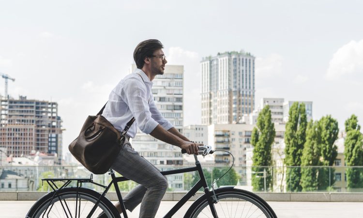The picture shows a cyclist in summer work clothes. The cyclist is carrying a large shoulder bag. He is riding his bike through the city.