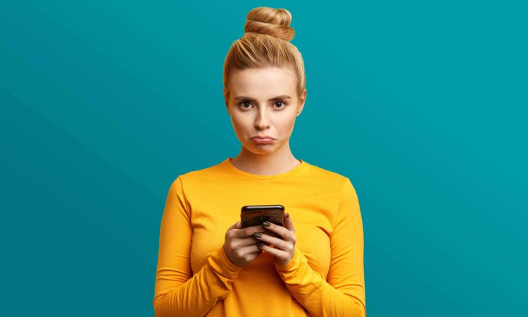 A young woman in a yellow shirt looks sadly at her hands, which she holds in front of her. She is standing in front of a light blue background.