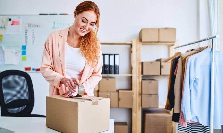 A woman with red hair stands in an office and prepares a parcel for the post office. Other parcels and items of clothing can be seen in the background.
