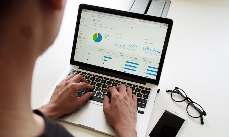 The picture shows a person at a laptop. The screen shows various diagrams and data on the financial management of a company. On the desk are a pair of glasses and a cell phone.