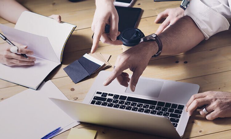 Mehrere Personen sitzen an einem Schreibtisch und zeigen auf einen Bildschirm.